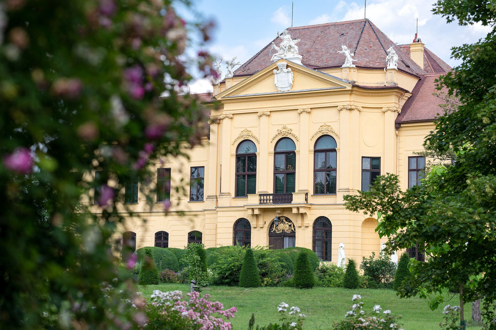 Blick auf die Fassade von Schloss Eckartsau. Im Vordergrund grüne Bäume und Blumen. 