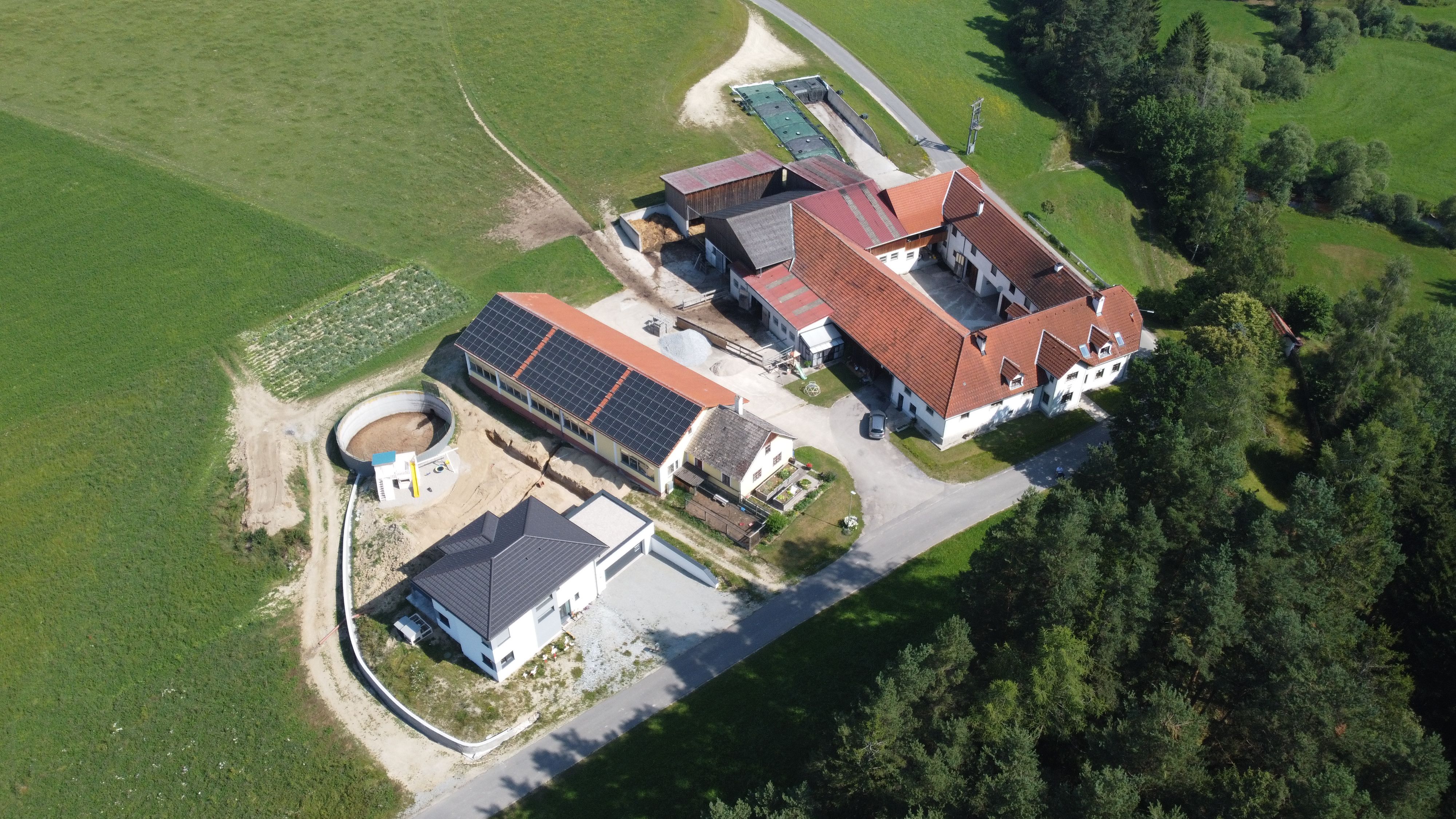 Luftaufnahme eines Bauernhofs mit mehreren Gebäuden und Solarpanelen auf einem Dach, umgeben von Feldern und Wäldern.
