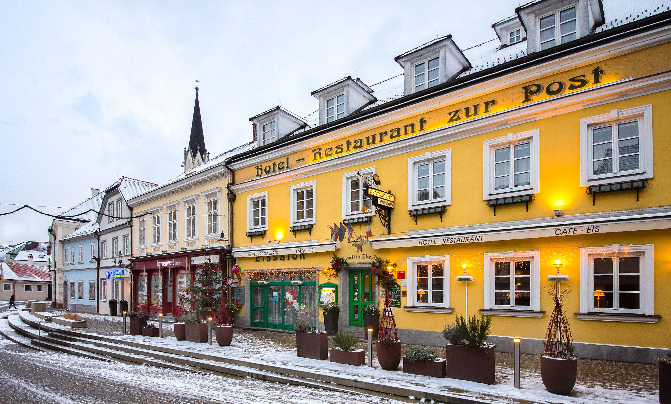Außenansicht eines gelben Gebäudes mit der Aufschrift 'Hotel-Restaurant zur Post' im Winter, umgeben von Schnee und dekoriert mit Lichtern.