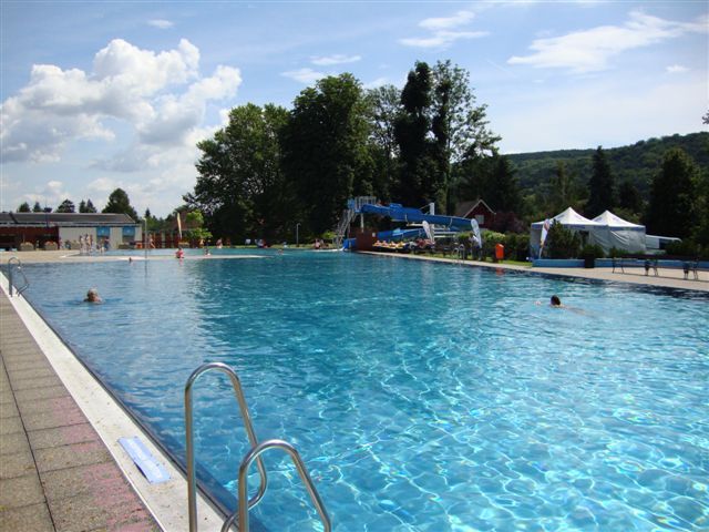 Ein großes Freibad mit klarem Wasser, umgeben von Bäumen und Hügeln unter einem blauen Himmel.