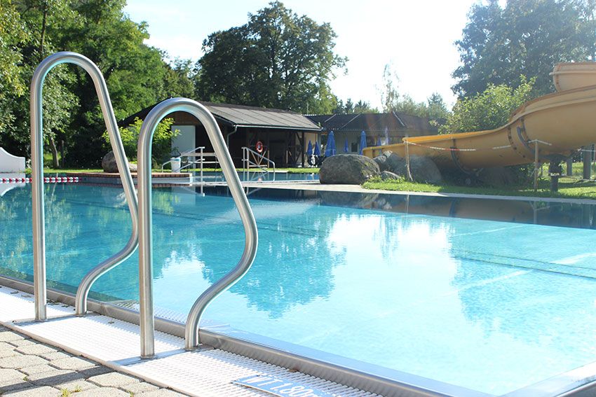 Ein Freibad mit Rutsche und Einstiegsleiter, umgeben von Bäumen und einem Gebäude im Hintergrund.