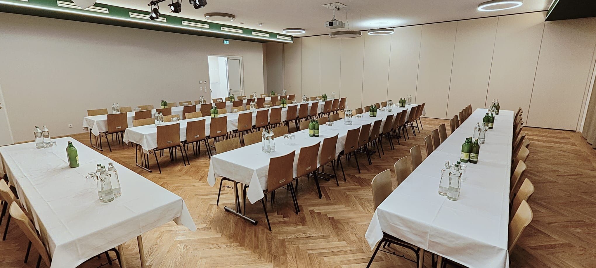 Großer Saal mit Holzstühlen, langen Tischen, weißen Tischdecken und Getränkeflaschen.