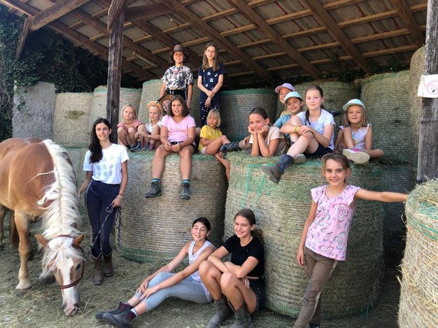 Eine Gruppe von Kindern sitzt auf Heuballen in einem Stall, neben ihnen steht ein Pferd. Die Kinder lächeln und tragen Freizeitkleidung.