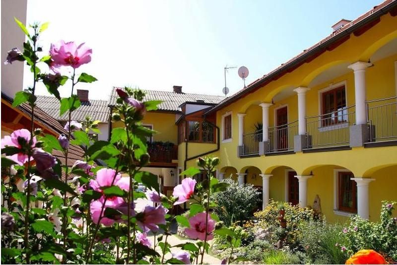 Ein sonniger Innenhof mit gelbem Gebäude, weißen Säulen und blühenden rosa Blumen im Vordergrund.