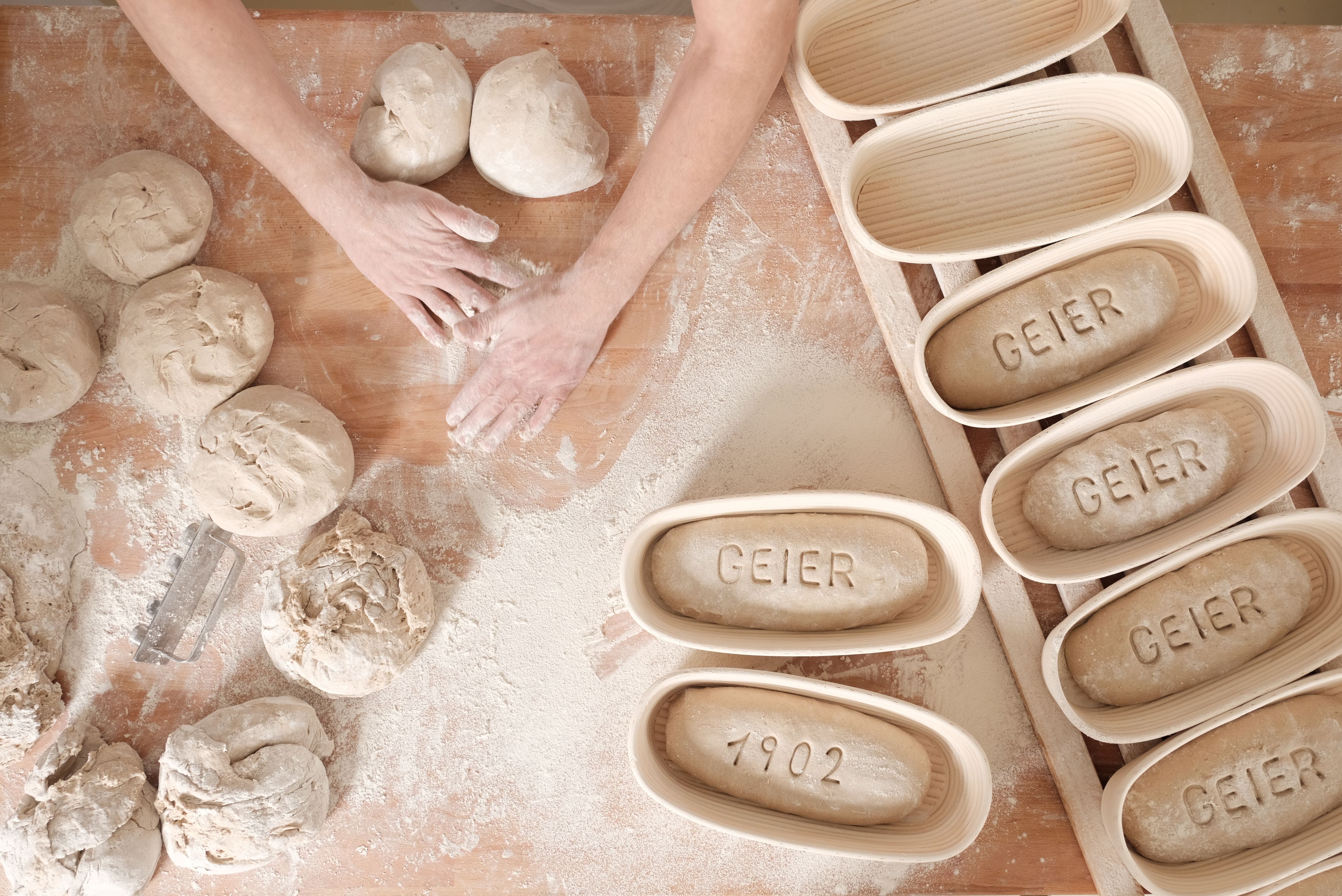 Hände formen Teig auf bemehltem Tisch, daneben Gärkörbe mit "GEIER" und "1902".
