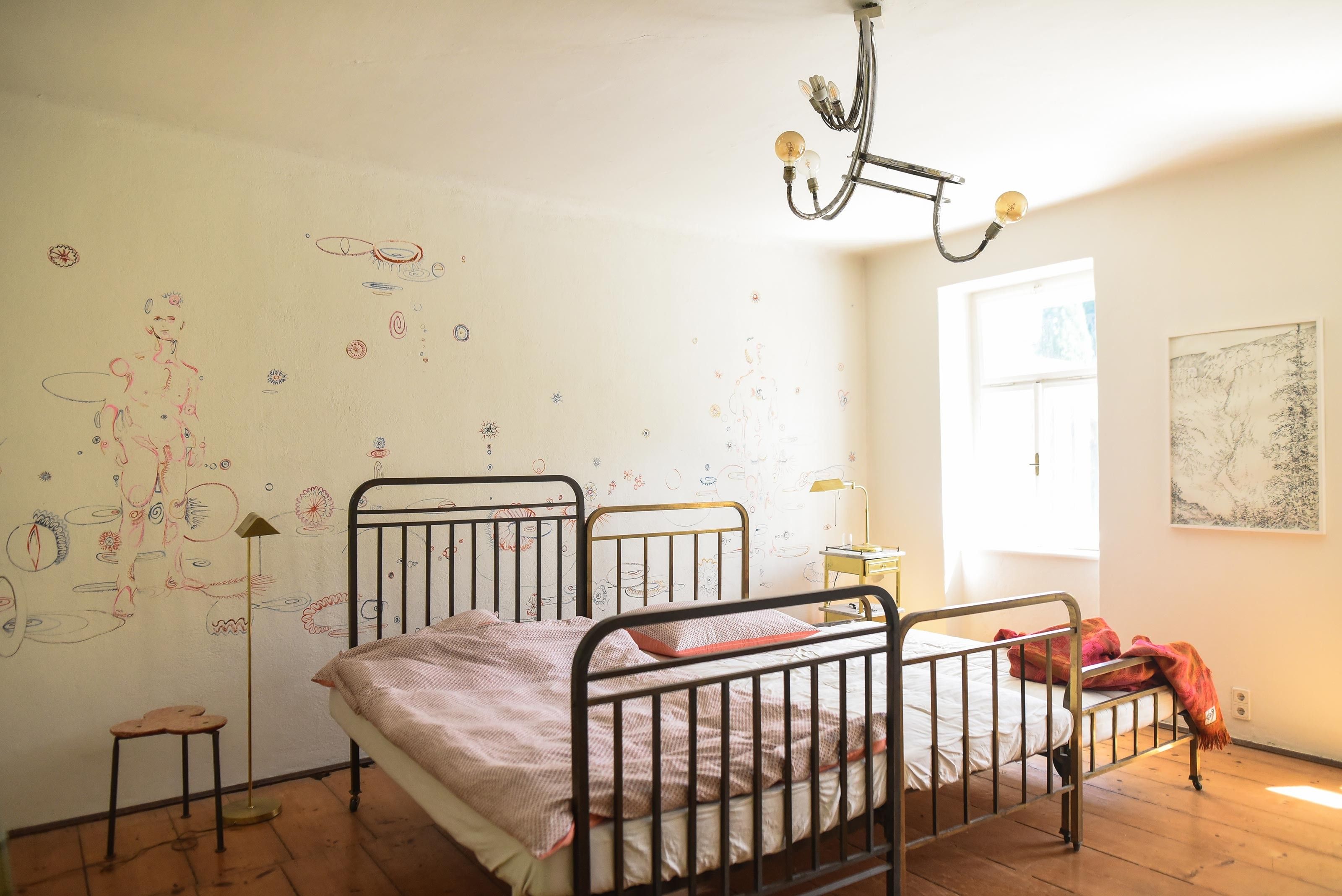 Ein helles Schlafzimmer mit einem Metallbett, Wandzeichnungen und einem Fenster, das Tageslicht hereinlässt.