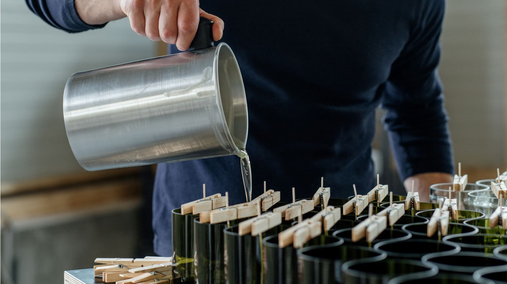 Person gießt Flüssigkeit aus einem Metallbehälter in Kerzenformen.