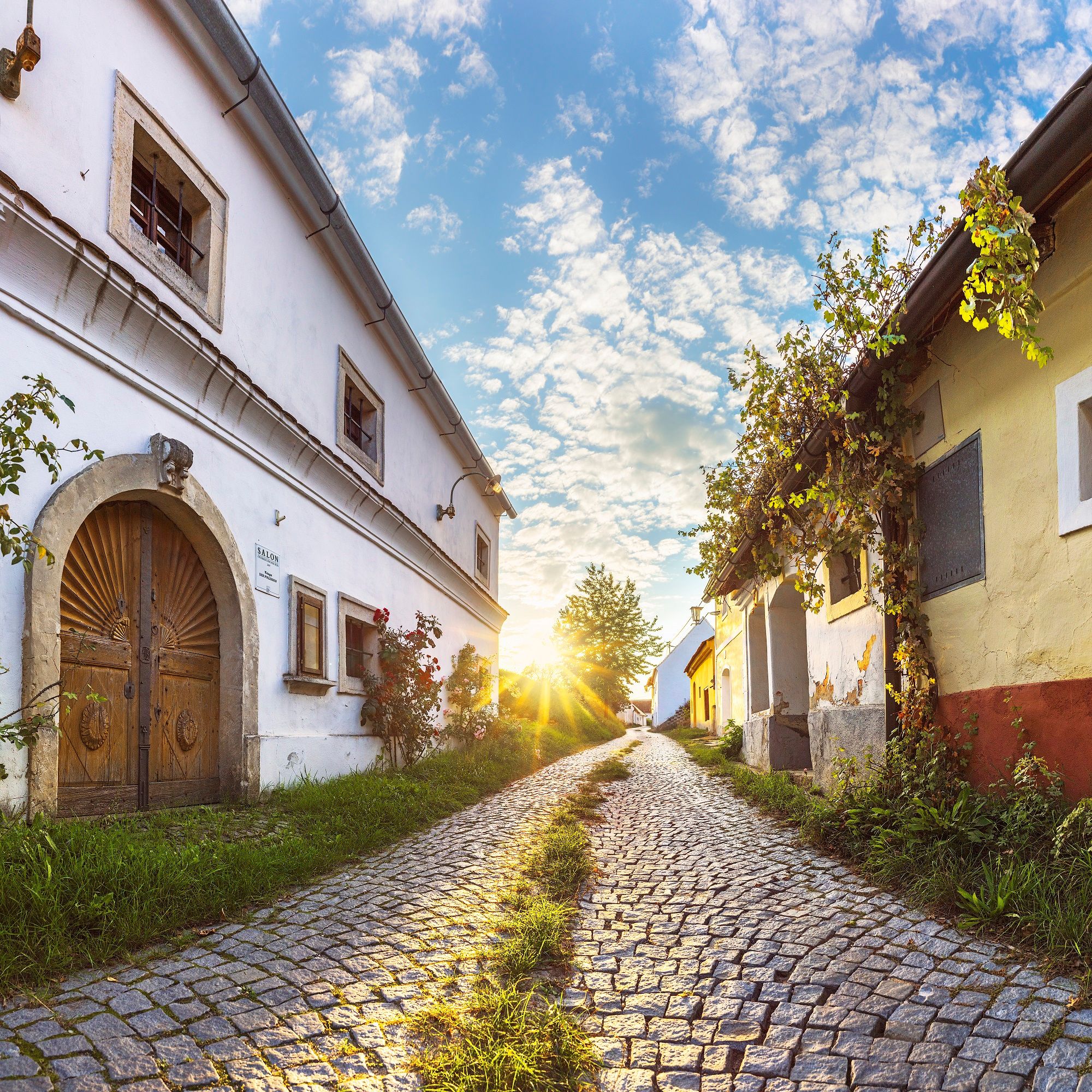 Kopfsteinpflasterstraße in Röschitz mit traditionellen Gebäuden und Sonnenuntergang im Hintergrund.