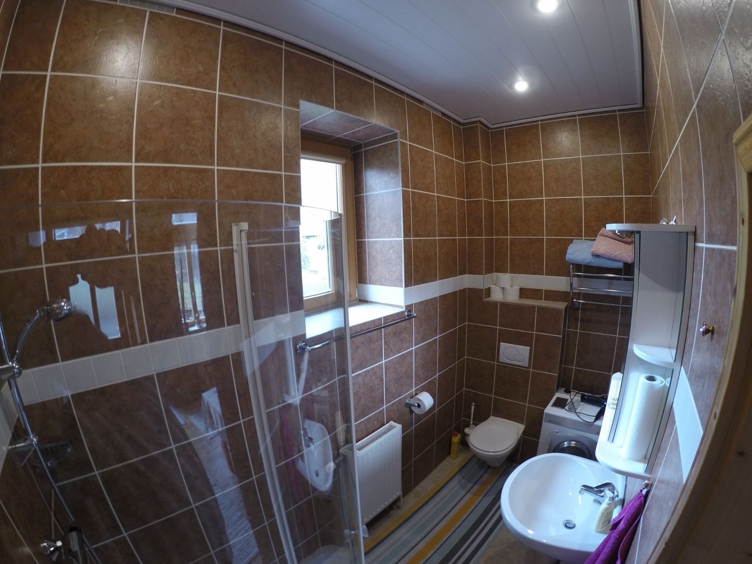 Badezimmer mit braunen Fliesen, Dusche, Waschbecken, Toilette und Waschmaschine.