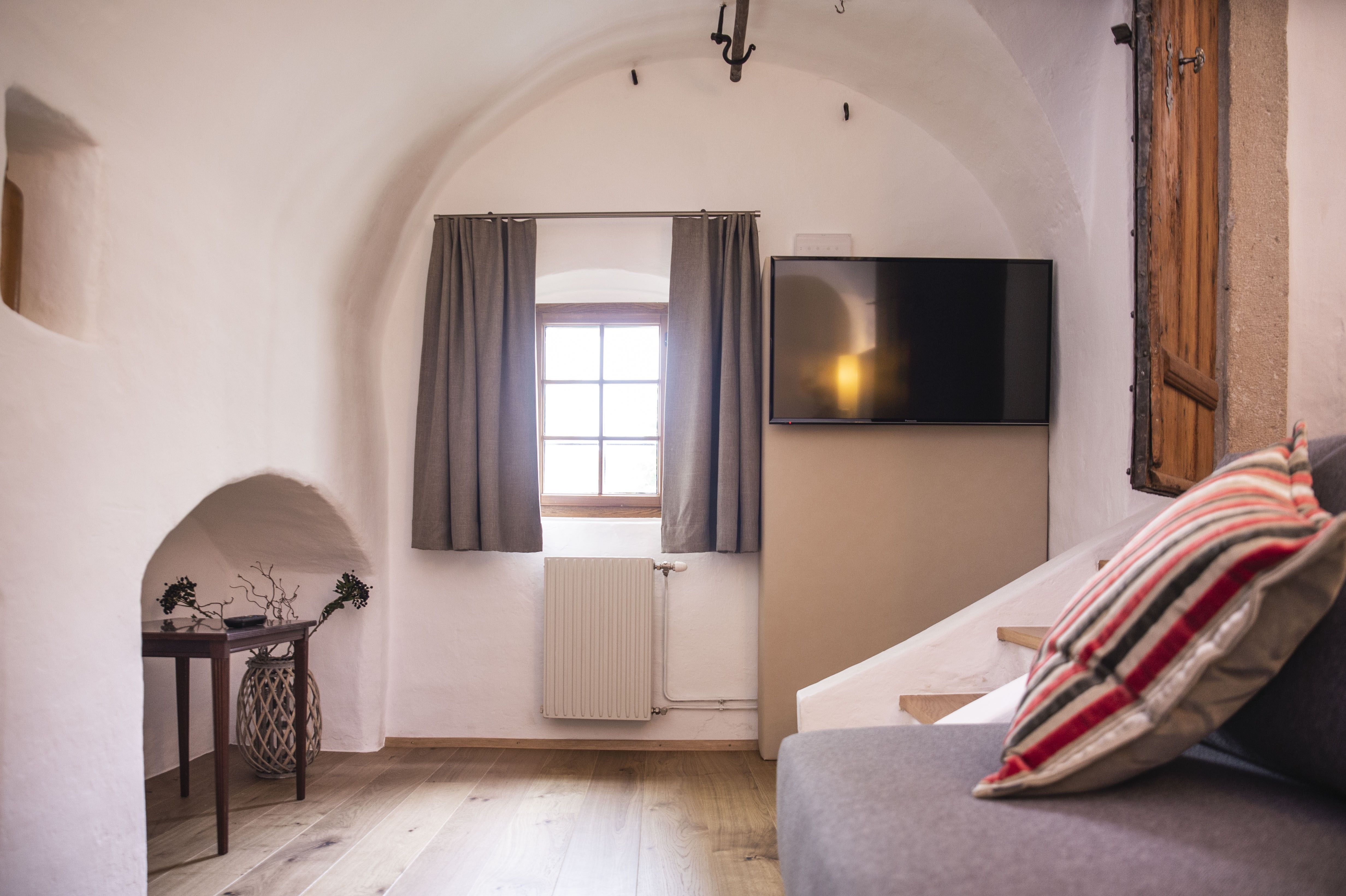 Innenansicht eines gemütlichen Zimmers mit gewölbter Decke, Fenster mit Vorhängen, Flachbildfernseher und Sofa mit gestreiften Kissen.