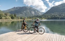 Die sanften Hügel und majestätischen Berge umrahmen den glitzernden See, während zwei Radfahrer auf dem Holzsteg innehalten und die atemberaubende Aussicht genießen. Die frische Luft und die ruhige Atmosphäre laden dazu ein, die Schönheit der Natur zu erkunden und unvergessliche Momente zu erleben.