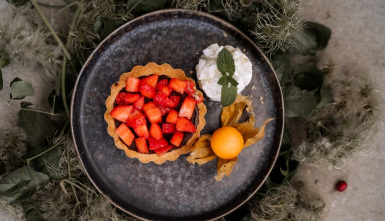 Ein Teller mit Erdbeert&ouml;rtchen, Sahne und Physalis, umgeben von gr&uuml;nen Bl&auml;ttern.