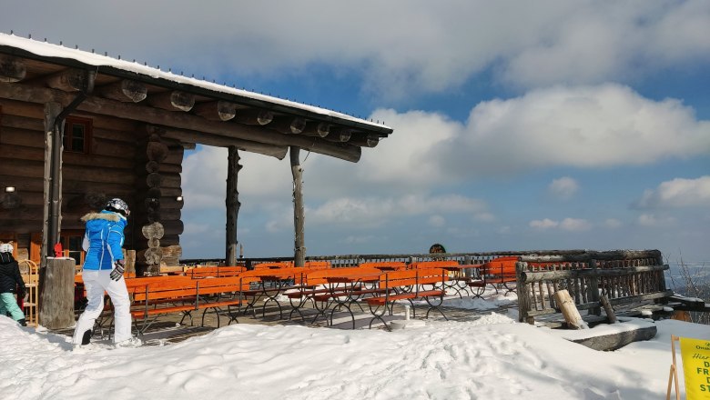 Verschneite Terrasse der Stoa Alm mit Holzb&auml;nken und Tischen, blauer Himmel und Wolken im Hintergrund.