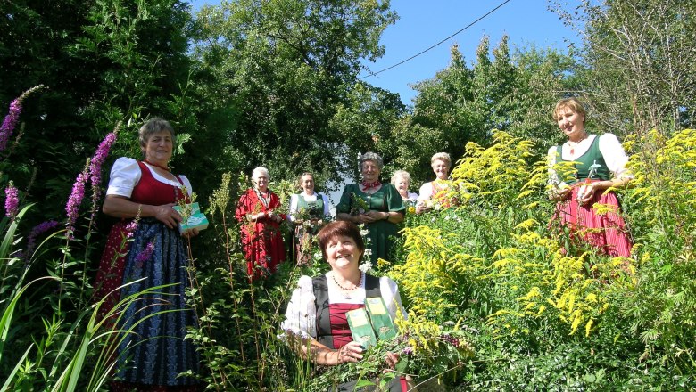 Frauen in traditioneller Kleidung stehen in einem Garten mit Kräutern und Blumen.