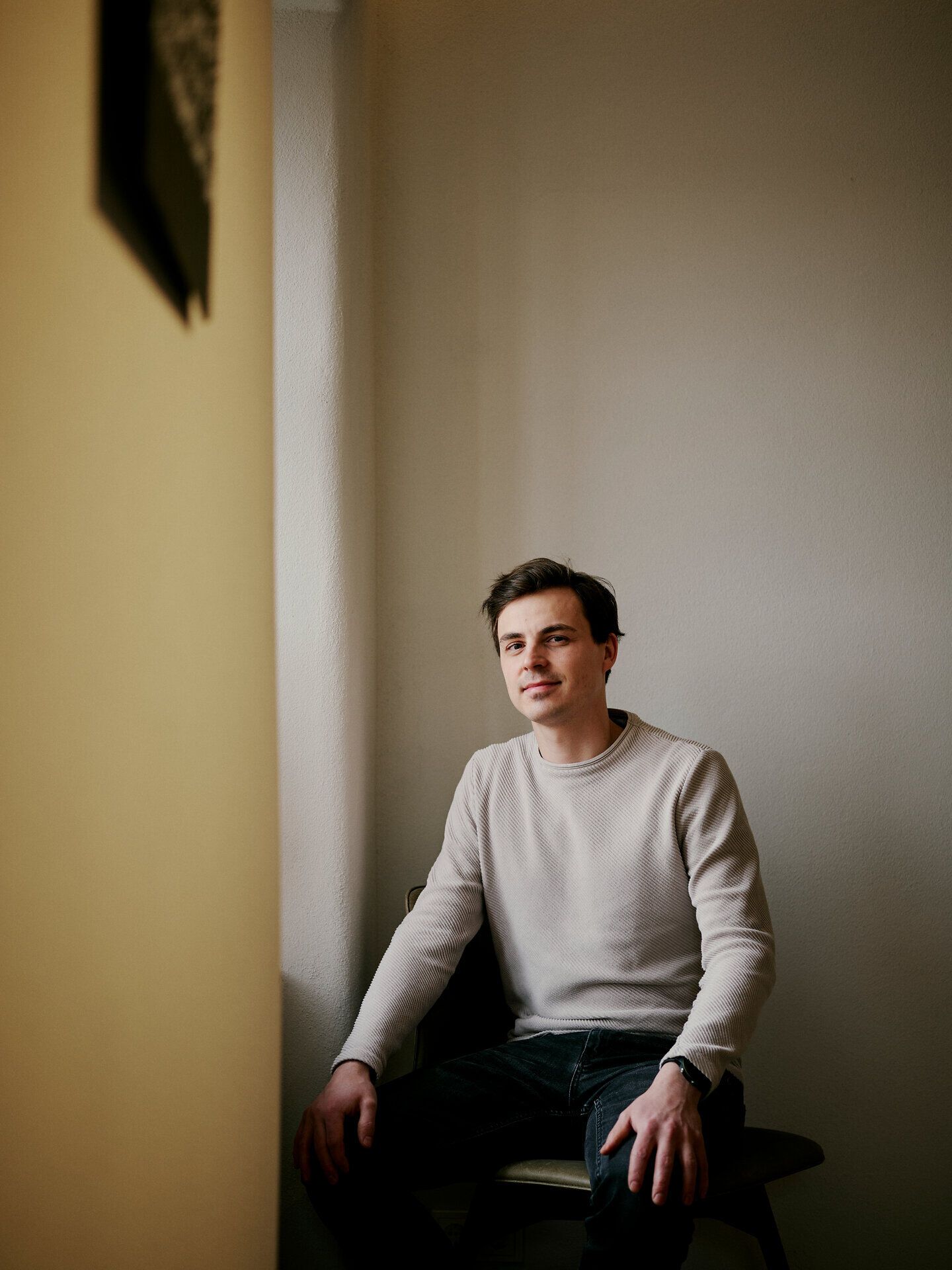 Eine Person sitzt auf einem Hocker in einem schlichten Innenraum und blickt in die Kamera