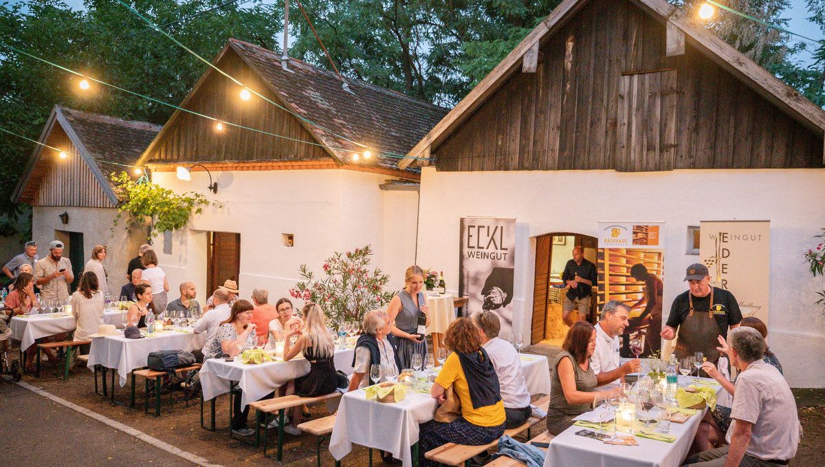 Menschen sitzen an Tischen vor einem Weingut in einer Kellergasse.
