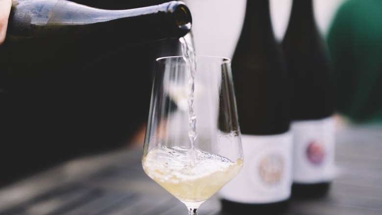 Wei&szlig;wein wird in ein Glas auf einem Tisch eingeschenkt, im Hintergrund stehen Weinflaschen.