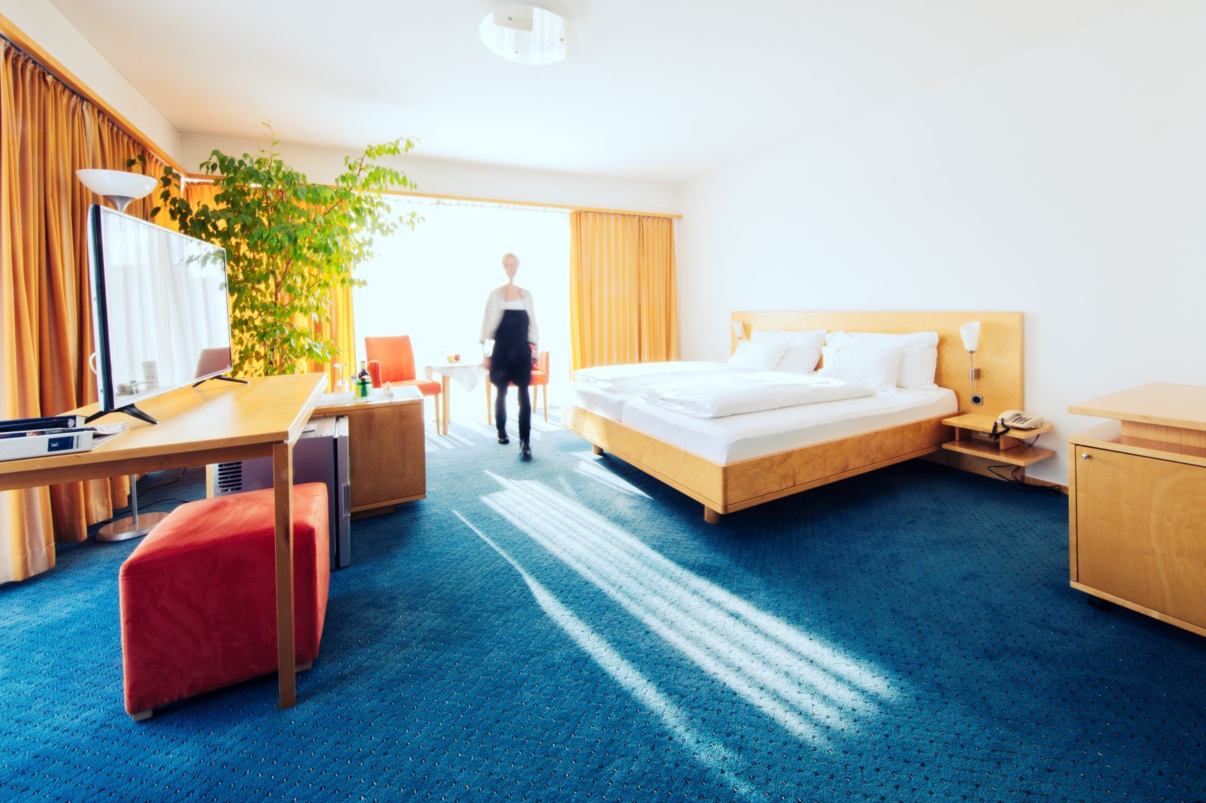 Helles Hotelzimmer mit Doppelbett, Schreibtisch, Fernseher und großer Pflanze. Eine Person geht durch den Raum.