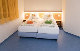 Ein Doppelzimmer mit zwei Einzelbetten, gelben Vorh&auml;ngen und blauer Boden. Die Betten sind mit Blumenbettw&auml;sche und farbigen Handt&uuml;chern dekoriert.