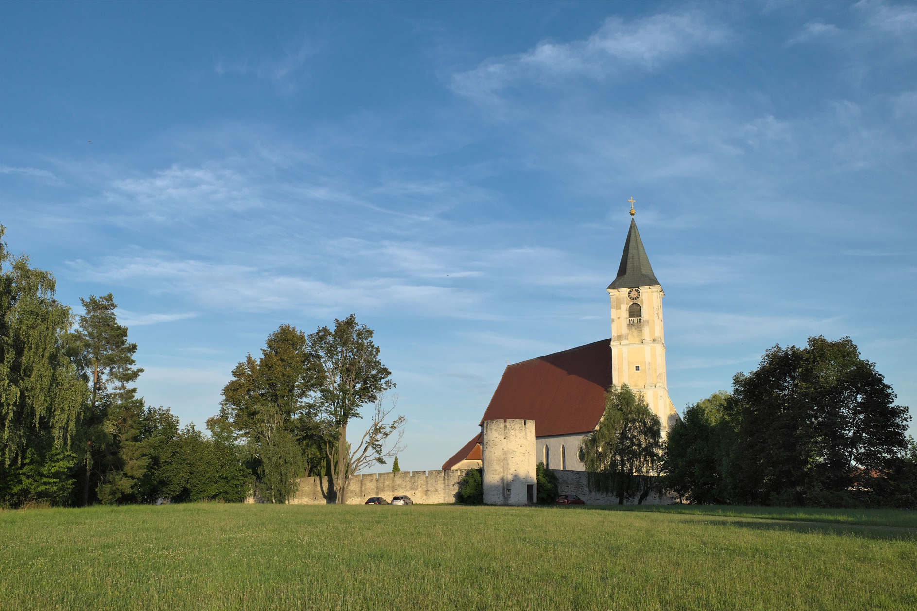 Kirche mit Turm und rotem Dach auf einer Wiese, umgeben von Bäumen und einer Mauer.