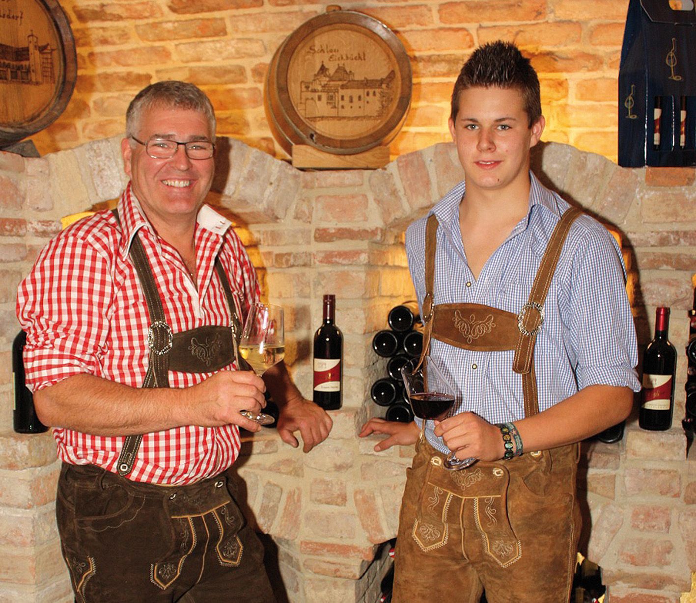 Zwei Männer in Tracht mit Weingläsern vor einer Ziegelwand.