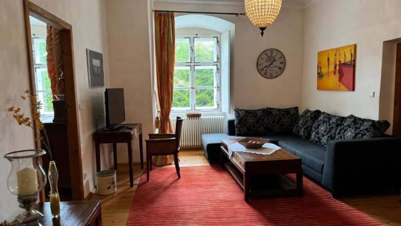 Gem&uuml;tliches Wohnzimmer mit Sofa, rotem Teppich, Kronleuchter und gro&szlig;em Fenster.