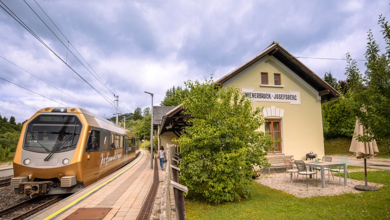 Urlaub am BAHNhof, © Niederösterreich Werbung / Maximilian Pawlikowsky Ein goldener Zug hält am Bahnhof Wienerbruck-Josefsberg, umgeben von grüner Landschaft und einem kleinen Sitzbereich im Freien.