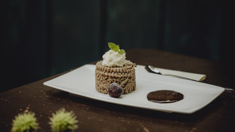 Ein Dessert aus Kastanienmousse auf einem weißen Teller, garniert mit Sahne und Minzblatt, daneben eine Schokoladensauce und eine Kirsche.