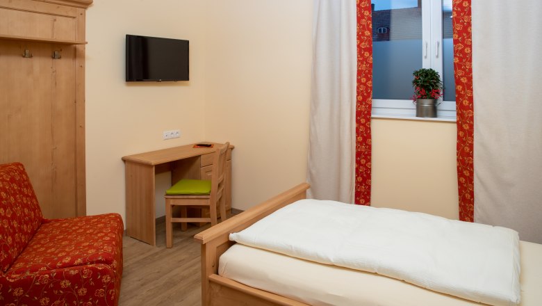 Ein gem&uuml;tliches Einzelzimmer mit Holzm&ouml;beln, einem Bett, einem Schreibtisch und einem Fernseher an der Wand. Ein Fenster mit Vorh&auml;ngen und einer Pflanze.