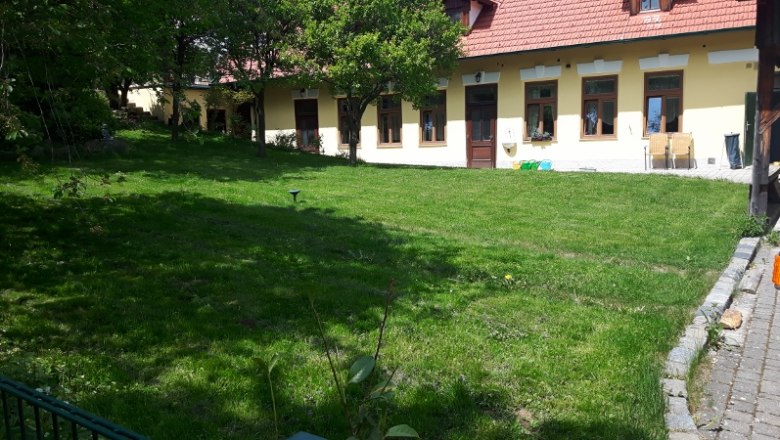 Gr&uuml;ner Garten vor einem gelben Geb&auml;ude mit roten Dachziegeln und mehreren Fenstern.