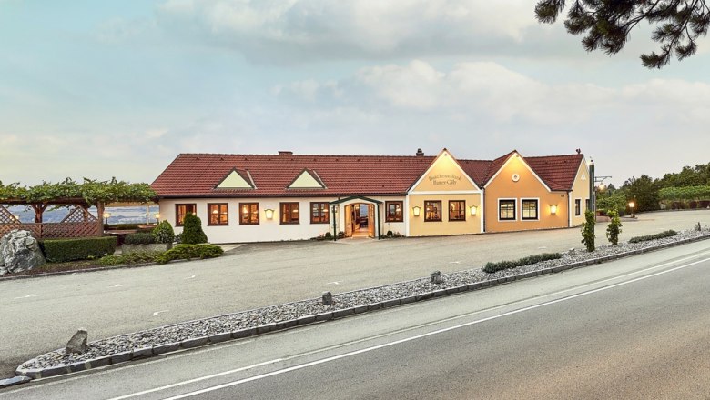 Weingut Humer - Heuriger, © Martin Sommer Außenansicht eines Weinguts mit gelbem Gebäude und rotem Dach, umgeben von Weinreben und einer Straße im Vordergrund.