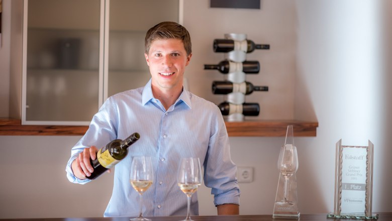 Ein junger Mann in einem blauen Hemd schenkt Wein in ein Glas ein, im Hintergrund Weinflaschen und eine Auszeichnung.