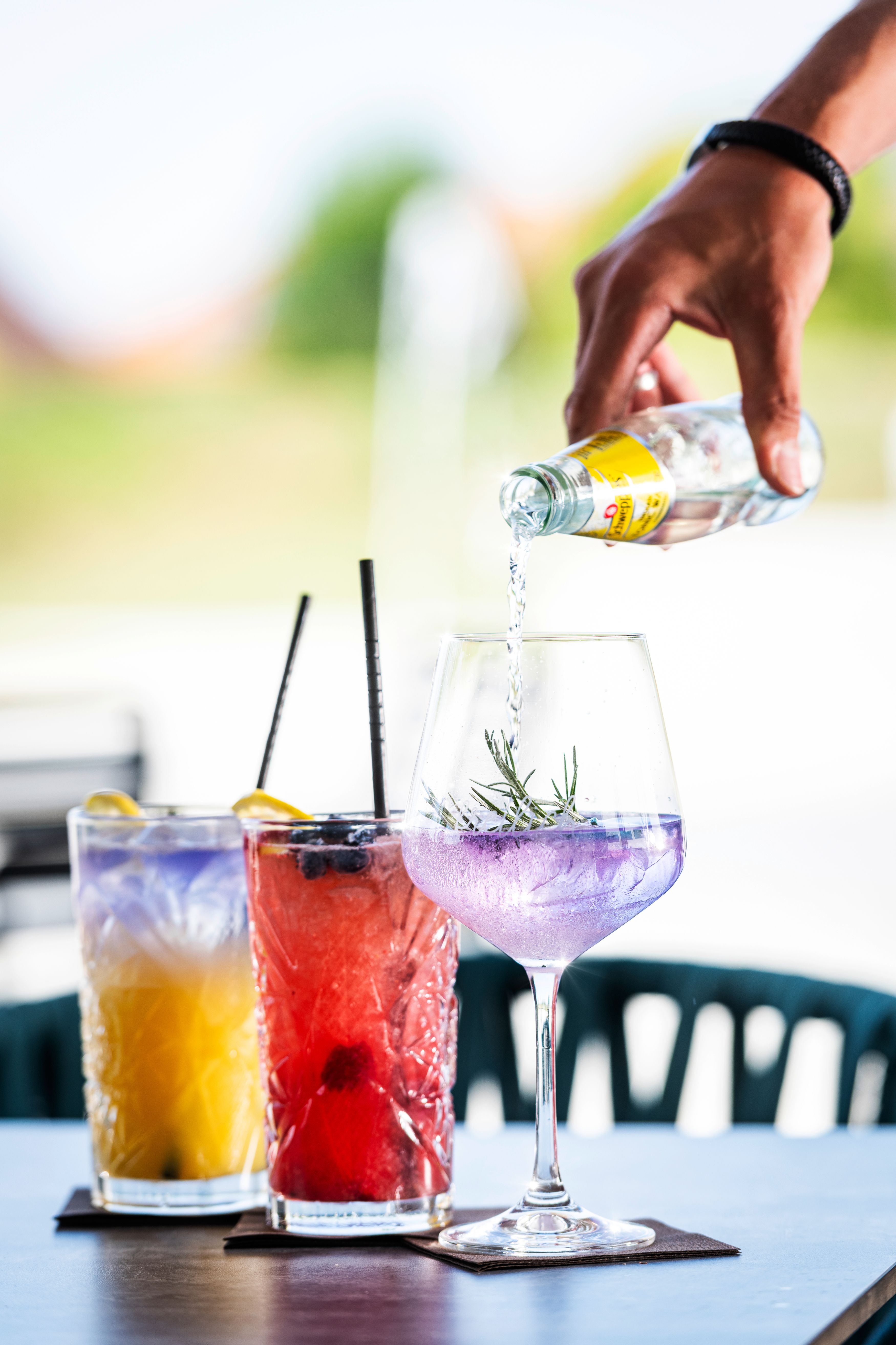 Drei bunte Cocktails auf einem Tisch, eine Hand gießt Flüssigkeit in ein Glas.
