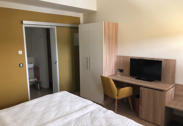 Ein modernes G&auml;stezimmer mit Bett, Schreibtisch, Stuhl, Fernseher und Schrank. Im Hintergrund ist ein Badezimmer zu sehen.