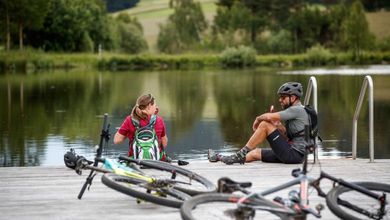 Zwei Radfahrer sitzen auf einem Steg am Frauenwieserteich, umgeben von Fahrr&auml;dern und Natur.