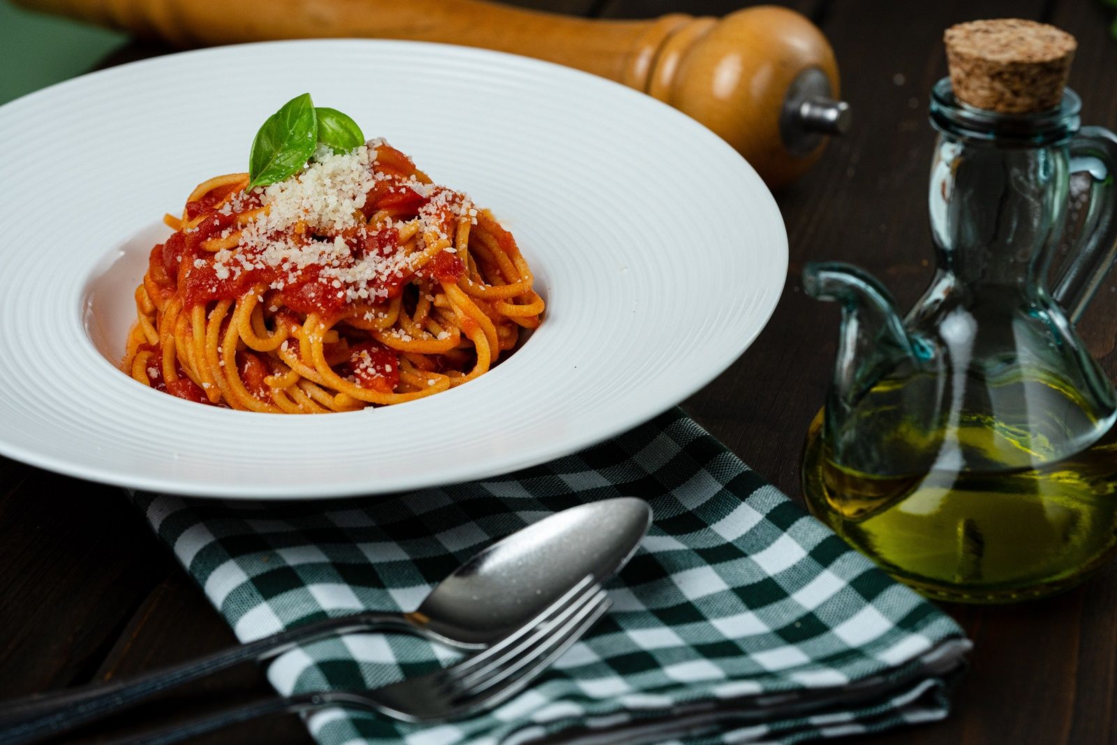 Ein Teller Spaghetti mit Tomatensauce, Basilikum und Parmesan, daneben eine Ölflasche und Besteck auf einem karierten Tuch.