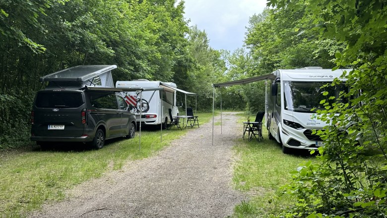 Zwei Wohnmobile und ein Van auf einem Campingplatz im Gr&uuml;nen mit ausgebreiteten Markisen und Campingst&uuml;hlen.