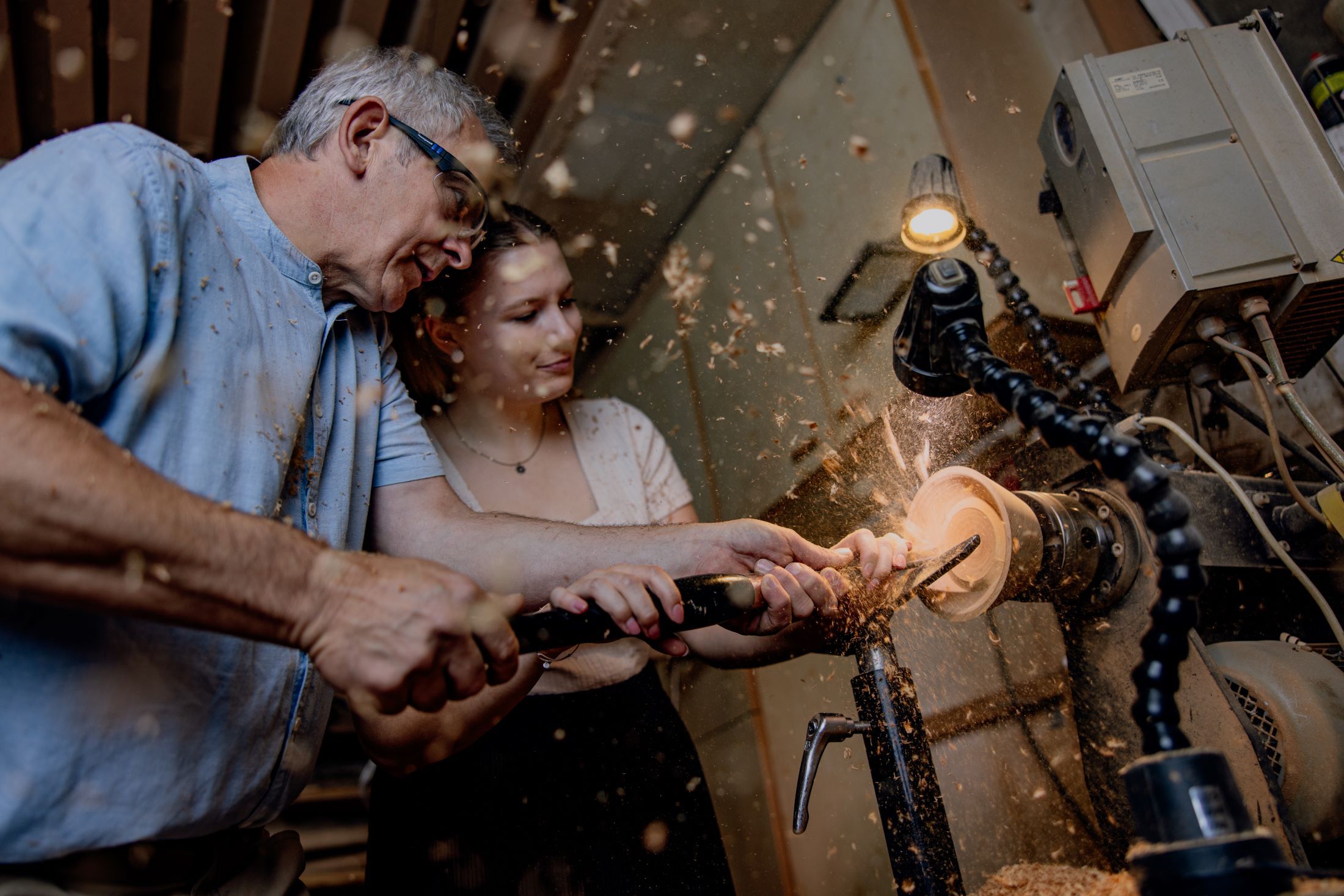Zwei Personen arbeiten gemeinsam an einer Drehbank, um Holz zu bearbeiten. Späne fliegen in der Luft.