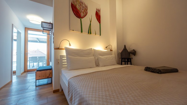 Modernes Schlafzimmer mit Doppelbett, heller Holzboden und Kunstwerk an der Wand. Tür führt zu einem Balkon mit Blick auf Dächer.