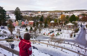 Winterlandschaft in den Kittenberger Erlebnisg&auml;rten mit schneebedeckten Wegen und bunten H&auml;usern im Hintergrund.
