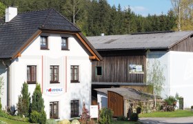 Gebäude der Mohnmühle Weinmann mit angrenzendem Holzschuppen, umgeben von Bäumen.