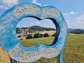Blick durch ein blaues Holzschild mit der Aufschrift 'Sonnige Gr&uuml;&szlig;e aus Hollenthon' auf eine l&auml;ndliche Landschaft mit H&uuml;geln und H&auml;usern.