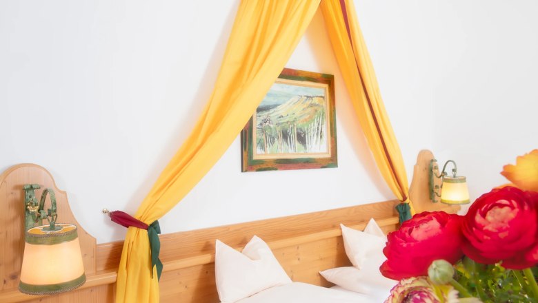 Ein gem&uuml;tliches Doppelzimmer mit gelbem Baldachin, Holzbett, Wandlampen und einem Blumenstrau&szlig; im Vordergrund.