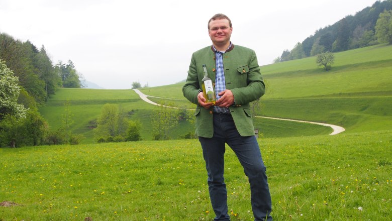 Mann in gr&uuml;ner Jacke h&auml;lt eine Flasche auf einer gr&uuml;nen Wiese mit H&uuml;geln im Hintergrund.