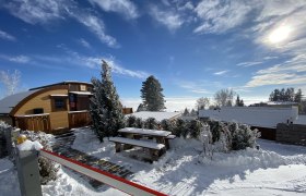 Winterliche Glamping-Hütte mit Schnee bedeckt, umgeben von Bäumen und blauem Himmel.