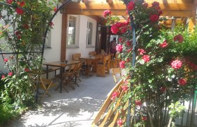 Ein Rosenbogen mit roten Rosen f&uuml;hrt zu einem Au&szlig;enbereich mit Holztischen und -st&uuml;hlen.