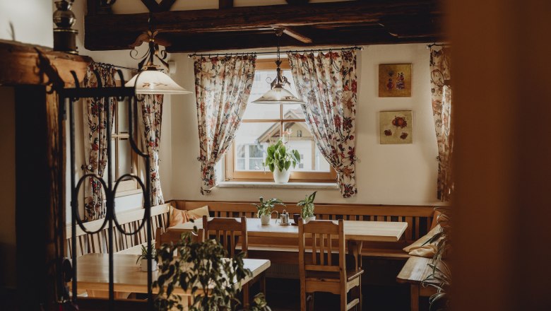 Gemütlicher Raum mit Holzmöbeln, Pflanzen und geblümten Vorhängen.