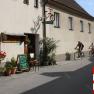 Außenansicht eines Hauses mit Fahrraddekoration und Weinausschank. Zwei Radfahrer fahren vorbei. Schild mit 'Zimmer frei'.