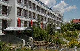 Modernes Gebäude mit mehreren Stockwerken, umgeben von grüner Landschaft und Fahrrädern im Vordergrund.