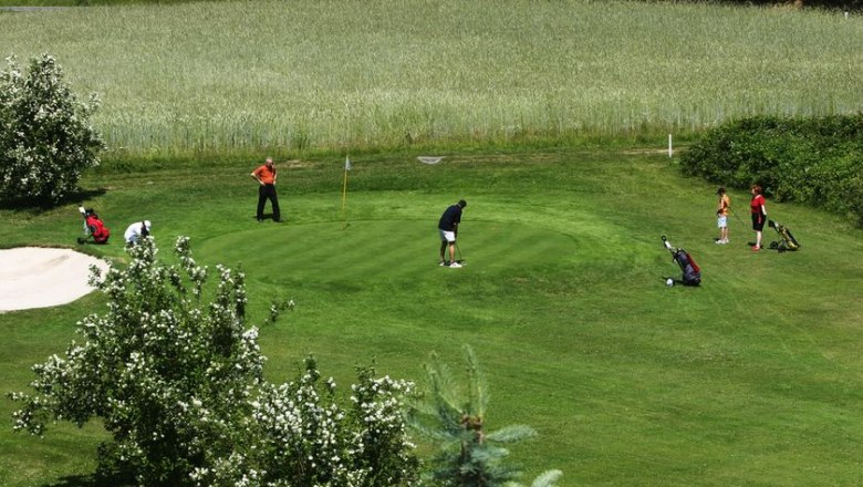 Menschen spielen Golf auf einem gr&uuml;nen Platz mit B&auml;umen und einem Sandbunker.